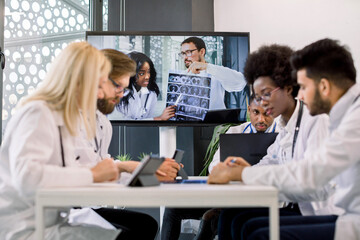 Multiracial doctors having a video conference at light office with two African and Caucasian doctors holding CT, sitting at the table and interacting each other, using tablets, laptop and making notes