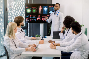 Naklejka premium Interracial group of doctors, scientists having discussion in meeting, while young African man telling new information about desease and treatment, showing results of research on the screen