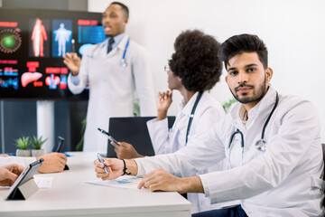 Portrait of young Indian male doctor, sitting at the table with his multiethnic colleagues, listening the speech of African man scientist, standing near big screen on the background