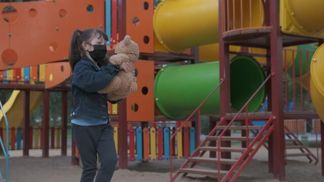 Play with bear on playground. A playful little girl jumps and spinnig with her bear toy bothe in protective mask on quarantined playground.