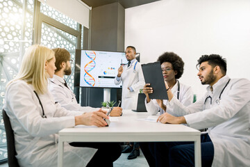 Fototapeta premium Multiethnic five doctors having a discussion during the meeting at conference room, using tablets and schemes. Handsome African male doctor speaker, showing biochemichal strusture