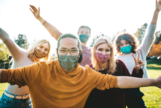 Happy friends covered by face masks having fun in the park - New normal concept with young people having party together outside.
