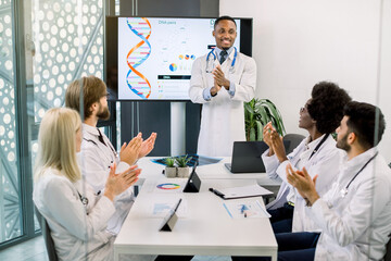 Multiethnic team of doctors applauding to cheerful handsome African male colleague, standing near the digital screen and having successful speech about DNA structure and new medicines