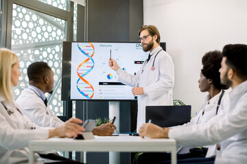 Fototapeta premium Handsome young Caucasian man doctor scientist wearing white coat standing near digital screen and giving presentation about biochemical molecules, new medicines to multiethnic group of his colleagues
