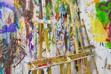 An easel, stained with paint, stands against the background of a painted wall. Art workshop.