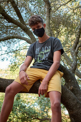 Naklejka na ściany i meble Young man sitting on a tree wearing a sanitary mask - concept of the new normal
