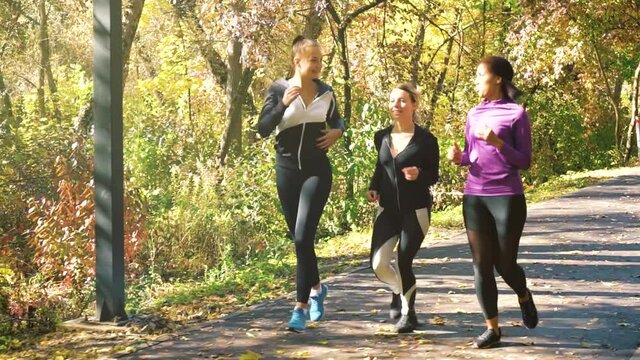 Young Women In Sportswear Jogging In Autumn Park, Yellow Trees On Background. Dolly Shot Multiracial Friends Running Together Outside. Concept Of Fitness