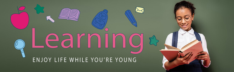 african american schoolgirl in glasses reading book near green chalkboard with learning, enjoy life while you are young lettering, panoramic shot