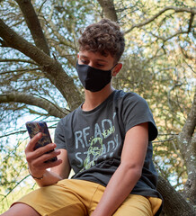 Young man sitting on a tree using his phone wearing a face mask-concept of the new normal