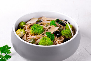 Pasta salad with whole grain spaghetti, romanesco cauliflower, olives, capers and parmigiano cheese in the grey ceramic bowl on the white wooden table