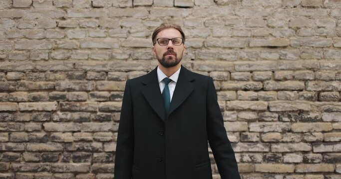 Portrait of handsome bearded man in black coat and suit posing on camera with brick wall behind. Businessman in eyeglasses standing outdoors and looking ahead.