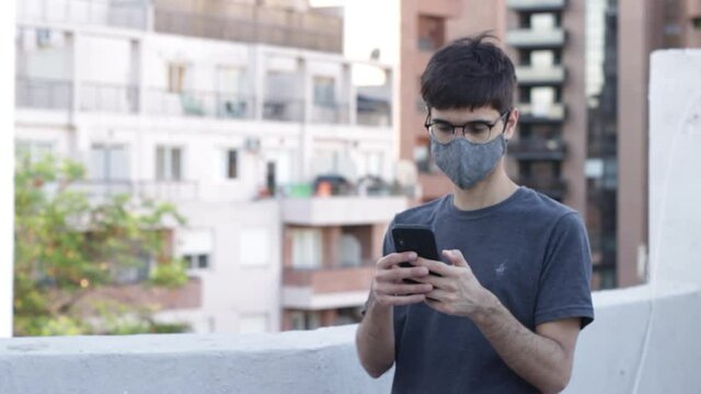 Young man using smartphone with a face mask during covid lockdown in a rooftop with city background