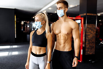 Fototapeta na wymiar Sportive young attractive couple of athletes, man and woman in medical protective mask on the face, show muscles in the gym, coronavirus, quarantine