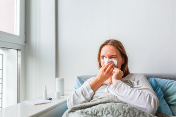 Sick woman blowing her nose with headache and fever lying under the blanket. Sick woman staying in bed with temperature durong coronavirus pandemic.