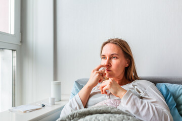 Sick woman spraying her nose with headache and fever lying under the blanket. Sick woman staying in bed with temperature durong coronavirus pandemic.