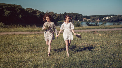 Two ladies running on the grass coast in the sunny morning summer country near the river or lake. Woman dressed in bright dress hold a bouquet of gathered wildflowers, nature background copy space.