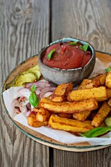 Fried potatoes (deep fried) with herbs and spices, ketchup, pickles and slices of pickled onion on a plate on a wooden background. Potato food . Baked potatoes with spices and salt.