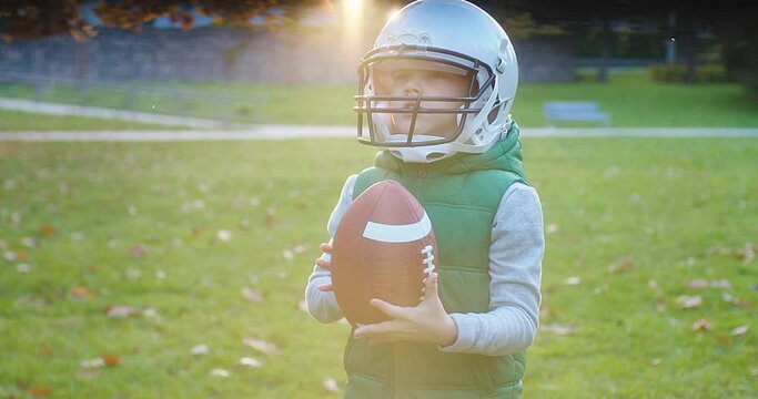 Cheerful happy child in helmet playing American football outdoors in sunny day at public park. Family sports weekend. 4K video.

