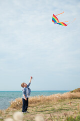 one active healthy small kid holding flying in air colorful kite standing on sea shore with blue sky on sunny summer holiday
