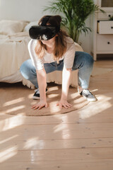 Naklejka premium Young woman in jeans and white T-shirt wearing virtual reality helmet plays game, works, watches video in room next to bed. Concept of modern technology, VR, augmented virtual games, entertainment.