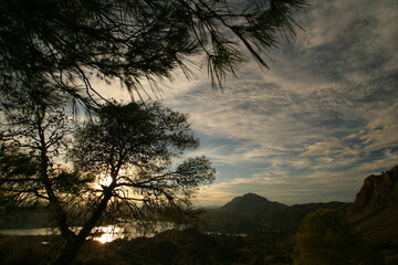 Árboles a contraluz, cielo con nubes, lago y montaña, al atardecer. Embalse del Quípar, Calasparra (Murcia).