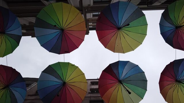Many colorful rainbow open umbrellas hang over narrow street between houses.