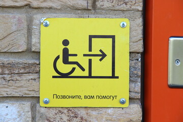 A yellow sign that reads in Russian 'Call, you will be helped' and with a picture of a person in a wheelchair, arrows and doors. The sign is attached by screws to a brick wall. 