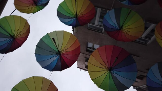 Many colorful rainbow open umbrellas hang over narrow street between houses.