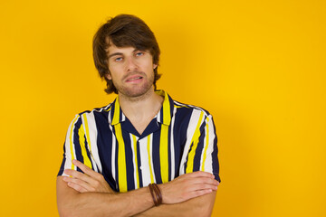 Young handsome Caucasian man, wearing stripped shirt standing against yellow wall bitting his mouth and looking worried and scared crossing arms, worry and doubt.