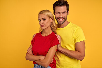 Naklejka premium Offended smiling young couple friends in colored t-shirts posing isolated on yellow background studio portrait. People lifestyle concept. Mock up copy space. Holding hands crossed, hugging.