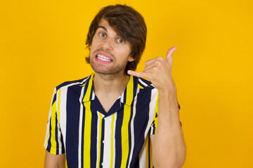 Young handsome Caucasian man, wearing stripped shirt standing against yellow wall imitates telephone conversation, makes phone call gesture with hands, has confident expression. Call me!