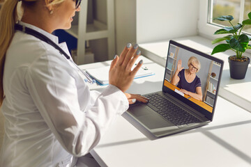 Doctor waving hand at laptop screen saying hello to senior woman during online consultation