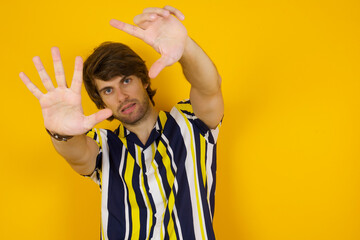Portrait of smiling Young handsome Caucasian man, wearing stripped shirt standing against yellow wall looking at camera and gesturing finger frame. Creativity and photography concept.