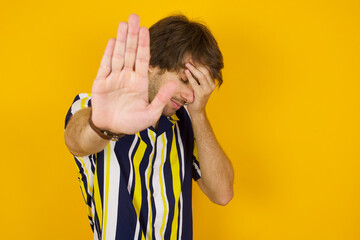 Young handsome Caucasian man, wearing stripped shirt covers eyes with palm and doing stop gesture,...