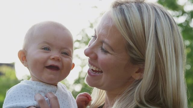 Loving mother cuddling and tickling baby daughter to make her laugh as she holds her outdoors in park or garden against flaring sun - shot in slow motion