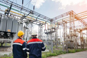 Two specialist electrical substation engineers inspect modern high-voltage equipment during sunset....