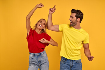 Funny young couple friends in colored t-shirts posing isolated on yellow background studio portrait. People lifestyle concept. Mock up copy space. Keeping eyes closed rising hands, dancing.