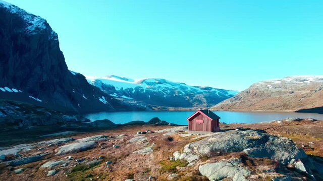 Small hut in the high mountains with a steep tall mountain slope wall and a lake in the background. 4K aerial video.