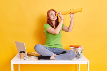 Red haired females casual attire sitting on table with crossed legs, making spyglass from paper and...