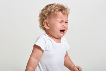 Little boy wearing t shirt cries, being in bad mood, looking away, posing isolated over white background, finding close person, having blond curly hair.