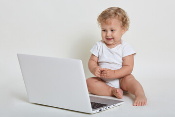 Charming blond curly haired infant wearing bodysuit, sitting barefoot on floor and looking directly at screen of lap top, barefoot toddler against white background.