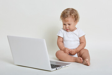Child with lap top computer sitting on floor isolated over white background, kid wearing bodysuit, posing barefoot with note book, looking cartoon with concentrated look.