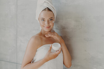 Indoor shot of happy young European woman applies anti aging cream on face, enjoys facial treatment...