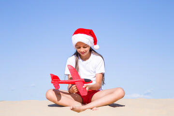 Christmas celebration vacations on a tropical beach: Happy cute little girl wearing a red Santa hat sitting on the sandy beach by the sea and holding a red toy plane on her hands on clear sunny day