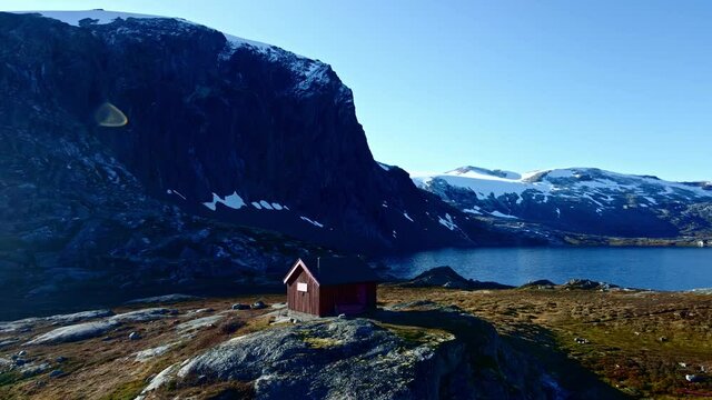 Small hut in the high mountains with a steep tall mountain slope wall and a lake in the background. 4K aerial video.