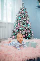 a baby boy in a striped jumpsuit is on all fours on a pink fur blanket with a gift and looks at the camera against the background of a Christmas tree. Vertical orientation