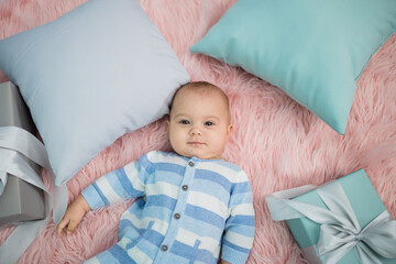 a happy little boy in a striped jumpsuit lies on his back on a pink fur blanket with gifts and pillows and looks at the camera