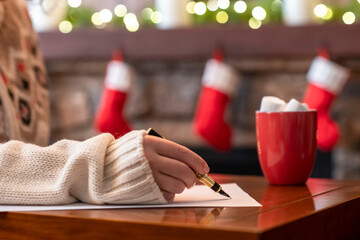 Woman writing letter to santa claus using fountain pen on sheet of paper at christmas fireplace with decoration of light bulbs drinking hot cocoa and marshmallow.