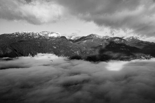Black and white image of a paraglider flying over a cloudy mountain valley
