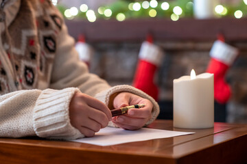 Obraz na płótnie Canvas Woman writing wish list using fountain pen on sheet of paper at christmas fireplace with decoration of light bulbs and candle on table.